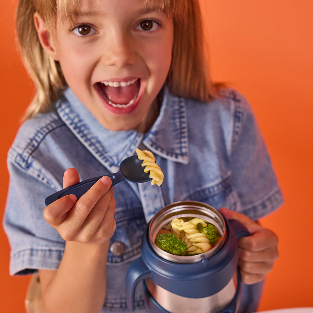 Child holding a bbox midnight colour thermos food jar with pasta and a spoon against an orange background
