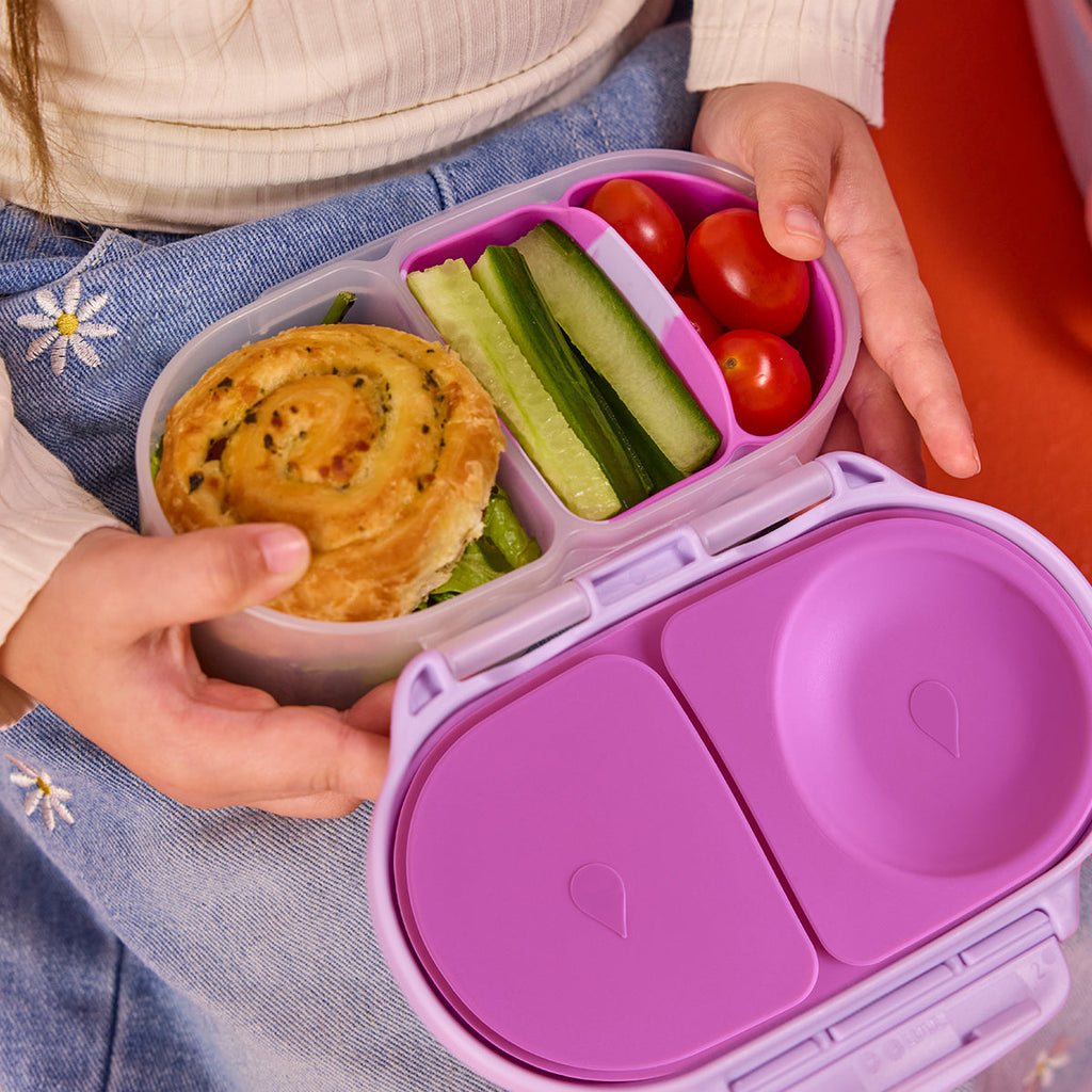 Purple bento box with compartments containing a pastry, vegetables, and tomatoes held by a person.