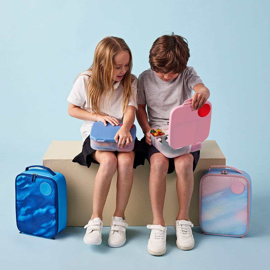 Two children sitting on a bench with bbox lunch boxes and colorful insulated lunch bags against a light blue background